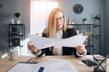 Confused mature lady with blond hair in eyeglasses sitting at table and holding papers in hands....
