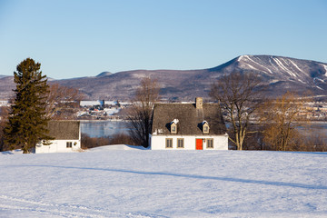 Beautiful early morning winter view of patrimonial white wooden house with steep shingled roof and shed in rural setting with trees, river and mountains, St. Pierre, Island of Orleans
