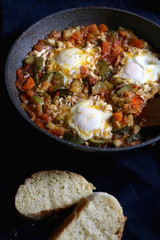 Shakshuka in a pan, with seasonal vegetables and eggs, and two slices of bread. Selective focus.