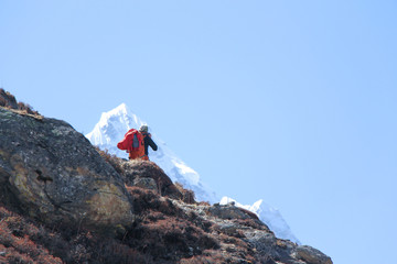 Back view. Photographer with orange backpack stands on mountainside in Himalayas and makes photo of...