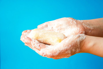 Fototapeta premium Women's hands are holding soap. Soap foam on the hands. Yellow soap in the hands. Woman washes soap with hands side view on a blue background. Virus protection. COVID-19