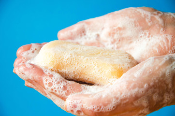 Women's hands are holding soap. Soap foam on the hands. Yellow soap in the hands. Woman washes soap with hands side view on a blue background. Virus protection.  COVID-19