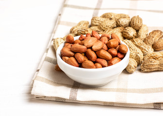 Peanuts in a shell and cleaned in a bowl on a checkered towel on a white wooden background. Copy space, close up