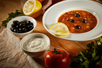 Macro Photo of Homemade Red Meat Soup with tomatoes, sausages, ham, beef, pickled cucumber, greens and vegetables on wooden background. Traditional pickle soup. Solyanka soup with olives and lemon.