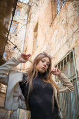 Street Fashion young attractive girl in a beige jacket, black turtleneck, black shiny pants and beige shoes against the background of urban locations