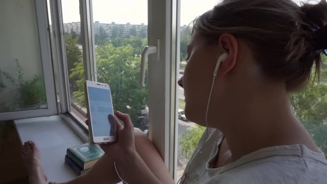 girl in home T-shirt, in white headphones, leafs through hand with list songs, files, videos on smartphone, listens music, rests her head against the window, sitting on the windowsill on balcony. In