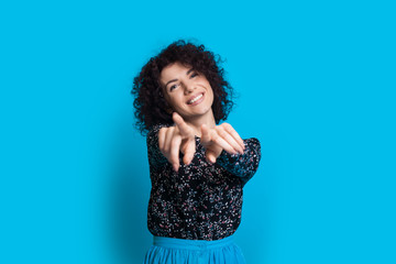 Joyous caucasian woman with curly hair is pointing to camera while posing on a blue background