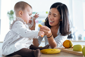 Happy young mother feeding her cute baby girl with a banana in the kitchen at home.