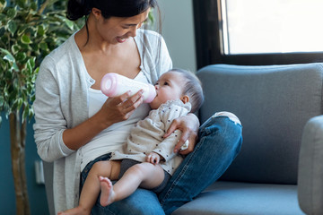 Happy young mother feeding her baby daughter with feeding bottle while sitting on sofa at home.