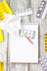 Notepad with medical supplies, thermometer and pills on white wooden background. Cold, virus flu...