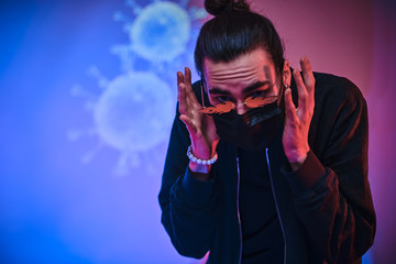 Young caucasian man wearing medical mask, sunglasses and black clothes looking scared and terrified. Isolated in a bright studio with coronavirus on the background
