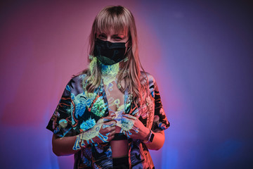 Sceptical looking young adult woman posin in a neon studio in front of wuhan coronavirus projection, wearing shirt, medical mask and sportive bra