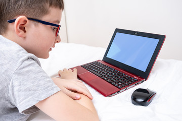 A boy wearing glasses lying in bed and using a laptop, typing the text on a keyboard close up, online education concept