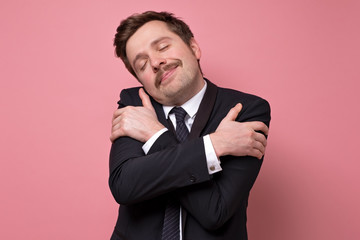 Handsome man wearing suit hugging oneself happy and positive, smiling confiden