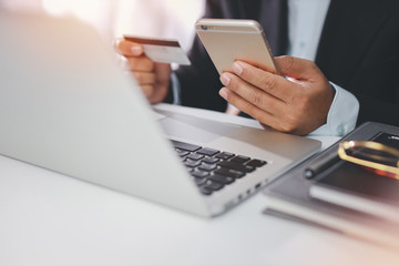 Man hands in black suit sitting and holding credit card and using laptop computer on table for online payment or shopping online. Business man using mobile phone while working. E-Banking concept