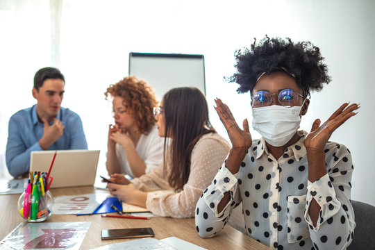 Serious businesswoman in protective mask looking at the camera in his office and people in background. Employee working in business office while wearing medical face mask