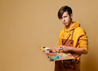 Young teen boy painter in yellow hoodie and jumpsuit standing and holding easel, paint and brush in...