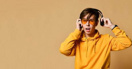 Young positive teen boy in yellow hoodie, sunglasses and headphones standing and looking at camera over yellow wall background