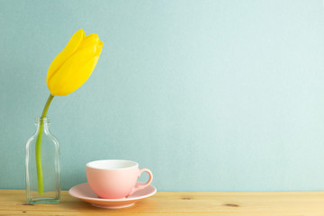 Coffee cup with yellow tulip flower in glass bottle on wooden table with mint green background. Floral arrangement, copy space