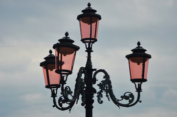 Fototapeta na wymiar The famous pink lights of Venice. Four lamps with pink glass are on an ornate black metal lamp post. The sky is overcast.