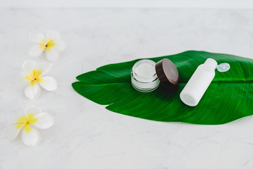 vegan natural beauty, pot of face cream and bottle of body lotion on tropical green leaf with monoi flowers