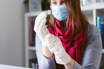 Young woman putting on protective gloves on hands wearing mask on face while working from home or...