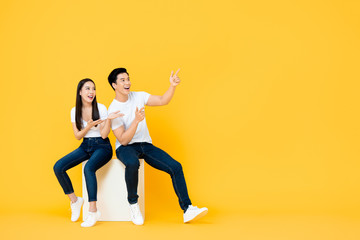 Fototapeta na wymiar Happy surprised attractive young Asian couple pointing and looking at blank space beside in yellow isolated stuidio background