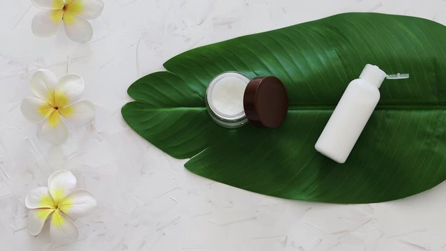 natural beauty without harsh chemicals, pot of face cream and bottle of body lotion on tropical green leaf with monoi flowers and camera panning vertically