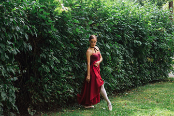 Woman ballerina in red ballet dress dancing in pointe shoes posing next to the wall of bushes