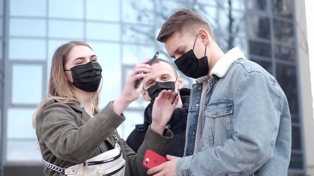Young people are near a modern scyscraper in black disposable masks.The boys and a girl are making selfies.