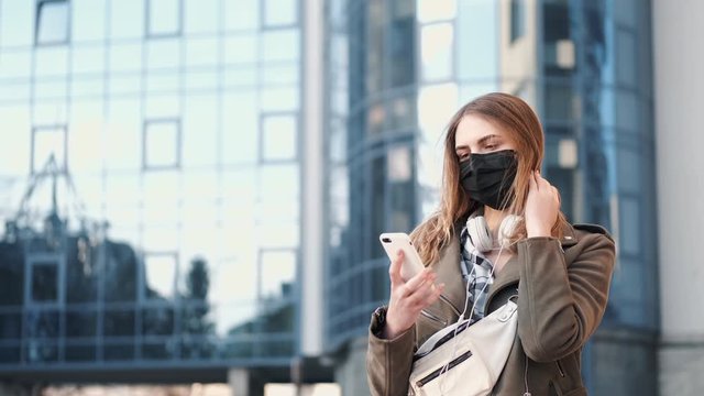 A young lady with a smartphone near a scyscraper is wearing black respirator.