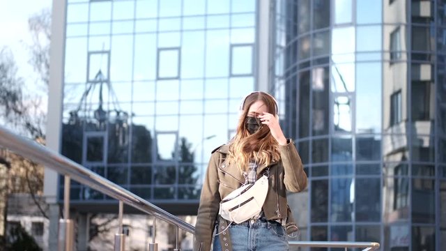 A young lady with a smartphone near a scyscraper is wearing black respirator.