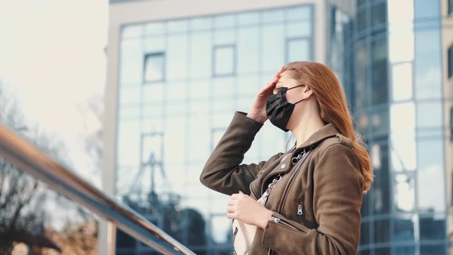 A young lady with a smartphone near a scyscraper is wearing black respirator.