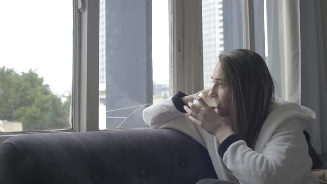 young woman sitting on sofa drinking coffee and staring out the window