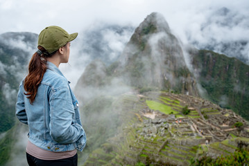 Young Asian woman traveler looking at Machu Picchu, one of seven wonders and famous tourist attraction in Cusco Region of Peru. This majestic place has known as 'Lost City of the Incas' - obrazy, fototapety, plakaty