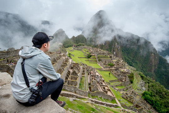 Young Asian man traveler and photographer looking at Machu Picchu, one of seven wonders and famous tourist attraction in Cusco Region of Peru. This majestic place has known as 'Lost City of the Incas' - Powered by Adobe