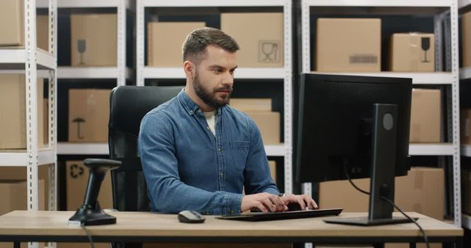 Handsome Caucasian young mailman sitting at desk in postal office store and working at computer. Male courier filling in invoice and entering data of parcel while typing on keyboard.