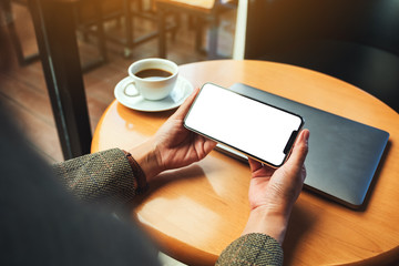 Mockup image of a woman holding mobile phone with blank white desktop screen with laptop and coffee cup on table