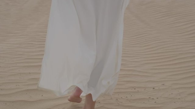 A brunette barefoot in a white dress fluttering in the wind walks along the desert sand. Slow motion.