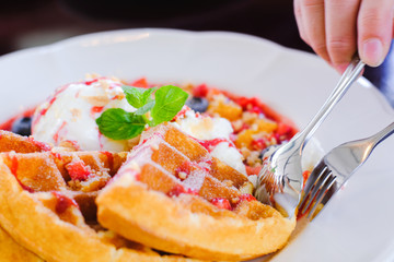 People hand used silver spoon and fork to eat Sweet waffles with strawberries sauce vanilla ice cream and fresh fruit on white plate at cafe restaurant in holiday summertime.