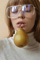 Fashion sensual young lady hold pear in mouth, stylish girl pretty face wear trendy glasses with fruit, vogue sexy attractive model in eyewear on beige studio background, vertical close up portrait