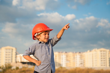 little kid builder in orange helmet pointing finger towards building on sunset and blue cloudy sky...