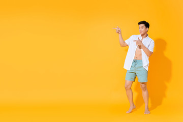 Full body of young handsome Asian man in casual summer outfit pointing hands to copy space aside on colorful yellow background