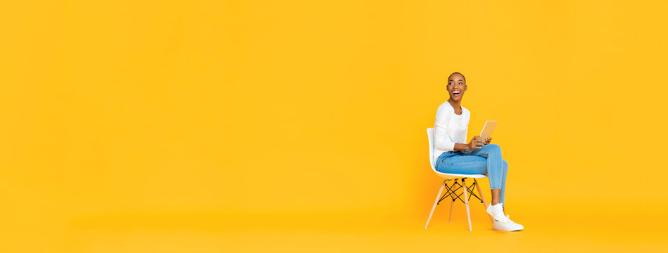 Trendy smiling African American woman sitting on a chair using tablet computer thinking and looking at empty space aside isolated yellow banner background