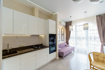 Kitchen room with a fresh renovation in beige tones. White kitchen set with kitchen appliances. semicircular window with curtains. TV, sofa and wicker furniture-table and two chairs