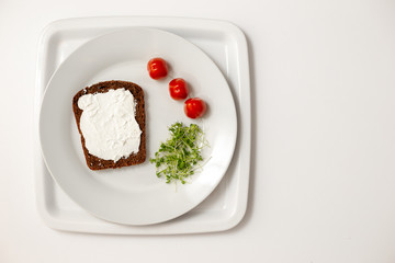 A slice of bread, spread with cottage cheese, lies on a white round plate, green watercress and red cherry tomatoes.