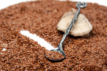 a light stone with a dark gray metal spoon on it lies against a background of brown flax seeds. On a spoon are flax seeds