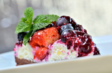 Delicious fruit strawberry and blueberry cream cheesecake tart glazed by cherry sauce and fresh mint leaves on decorative white plate. Low depth of field.