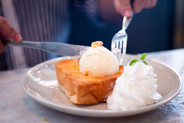 Hand of woman using knife and fork to eat honey toast with vanilla ice-cream and whipped cream at cafe