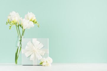 Festive spring background with standing gift box with ribbon, knot, white flowers freesia in glass vase in green mint menthe interior on white wood board.